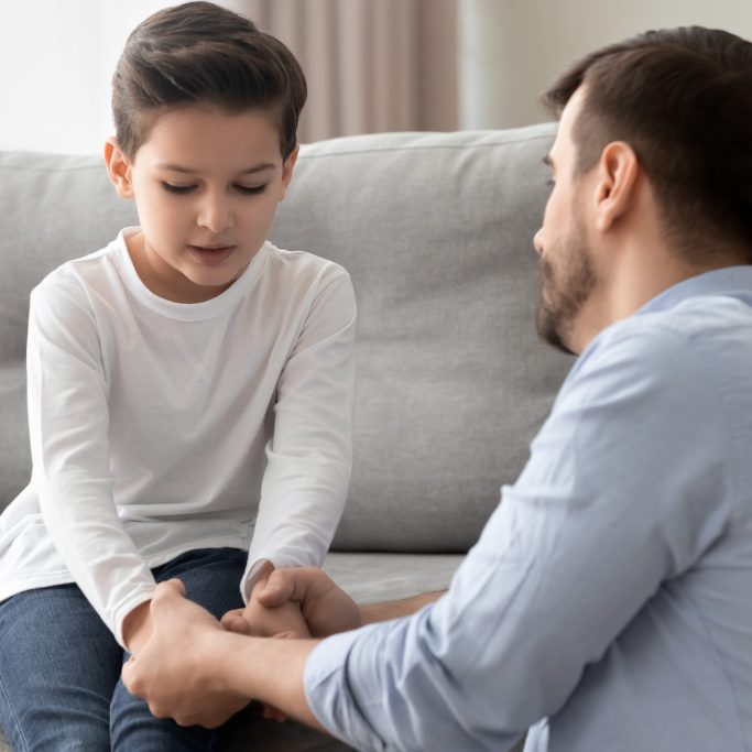 Worried loving young single father holding hand talking comforting upset little kid son sharing helping with problem, caring dad foster parent give support apologizing supporting listening child boy